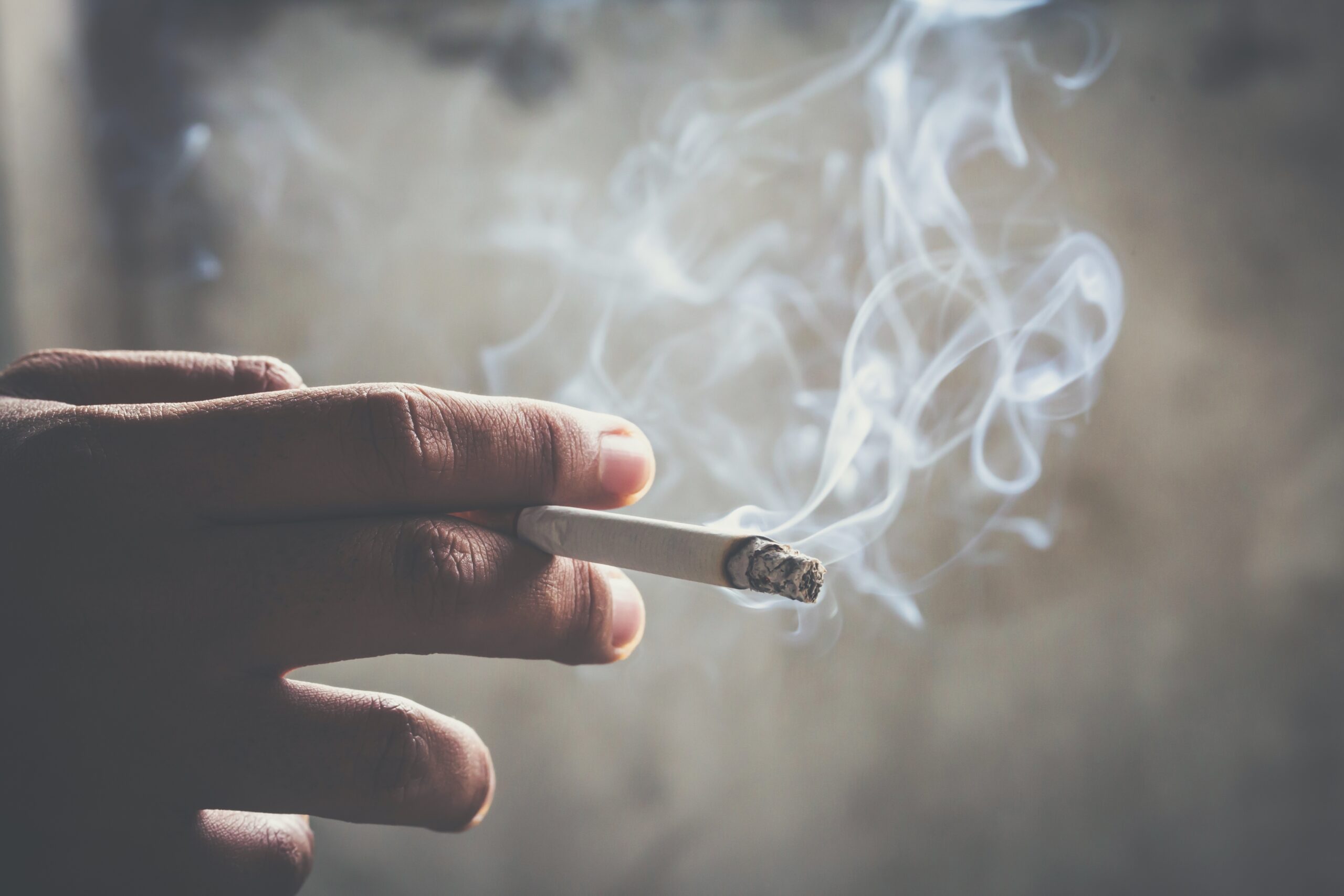 A hand holds a lit cigarette wafting smoke.