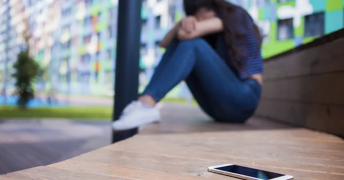 A distressed female teenage victim of social media harm next to a smartphone